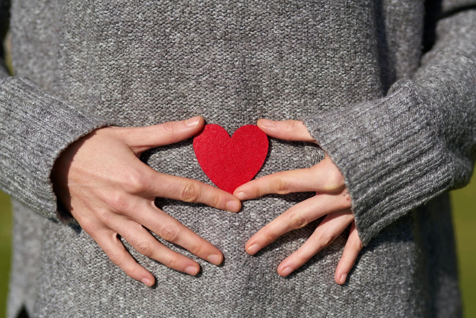 Hands in a grey sweater holding a red paper heart, symbolizing love and care.